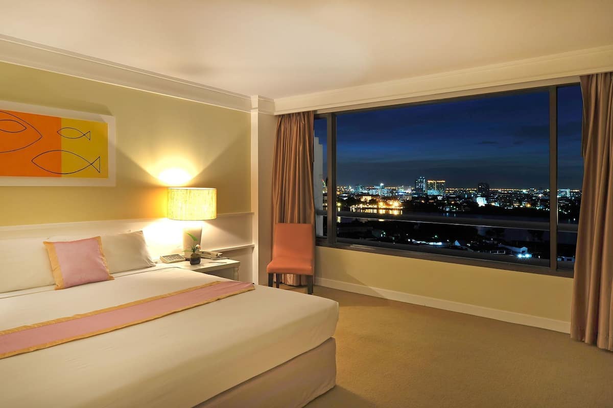 Une chambre d’hôtel comprenant un lit, une table de chevet, une lampe, une chaise et une grande fenêtre donnant sur la ville.