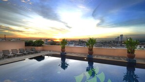 A rooftop pool with lounge chairs and potted plants, overlooking a cityscape at sunset.