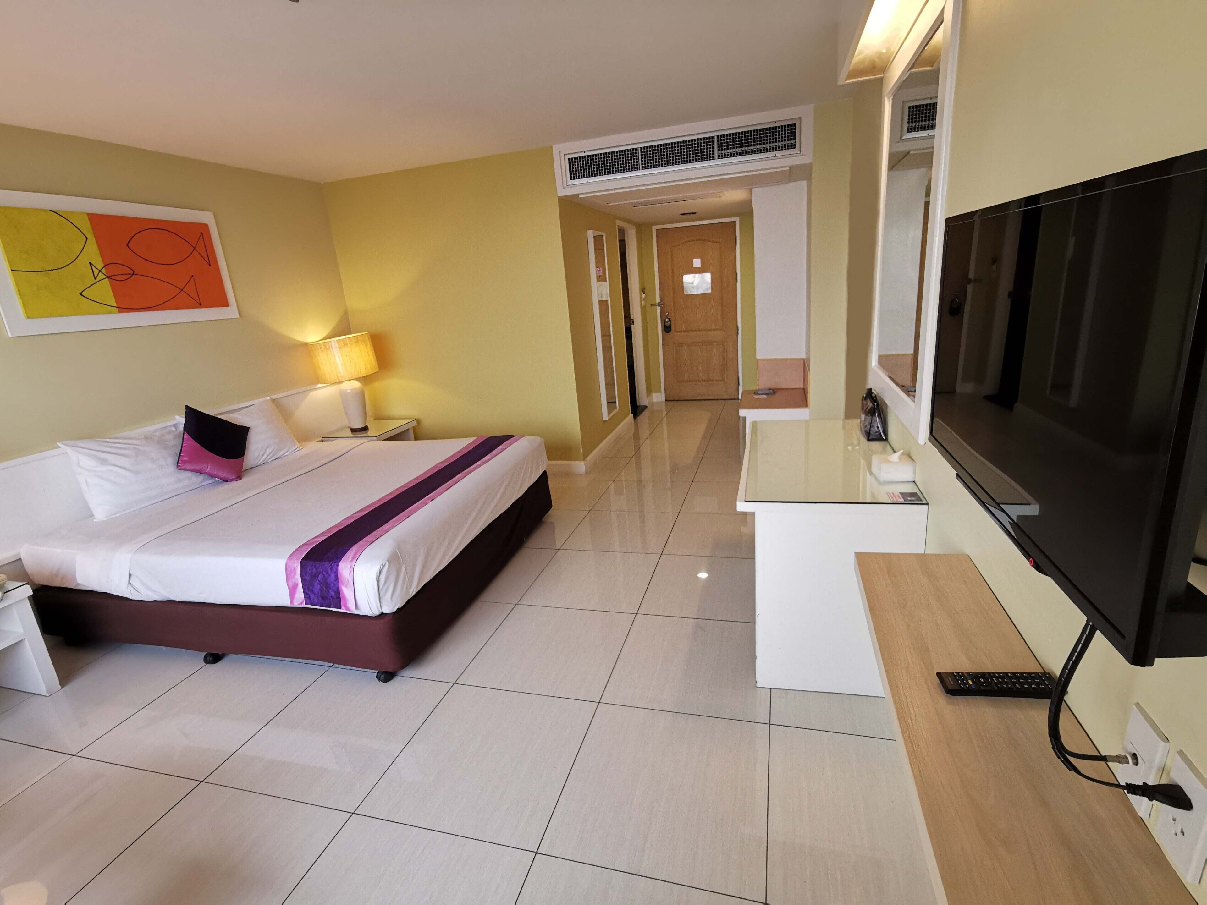 A hotel room with a bed, bedside table, lamp, TV, and a hallway leading to a door.
