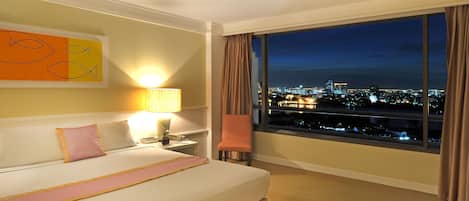 A hotel room with a large bed, a nightstand, a lamp, a chair, and a view of a cityscape at night.