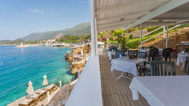Un restaurante frente al mar con terraza al aire libre, escalones de piedra y vistas a barcos y colinas.
