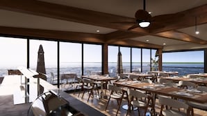 Un restaurante con vistas al mar, con mesas y sillas de madera, un ventilador de techo y amplios ventanales.