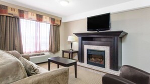 A living room with a fireplace, a flat-screen TV mounted above it, a sofa, a small table, and a lamp. - Ramada by Wyndham Pincher Creek (Pincher Creek)