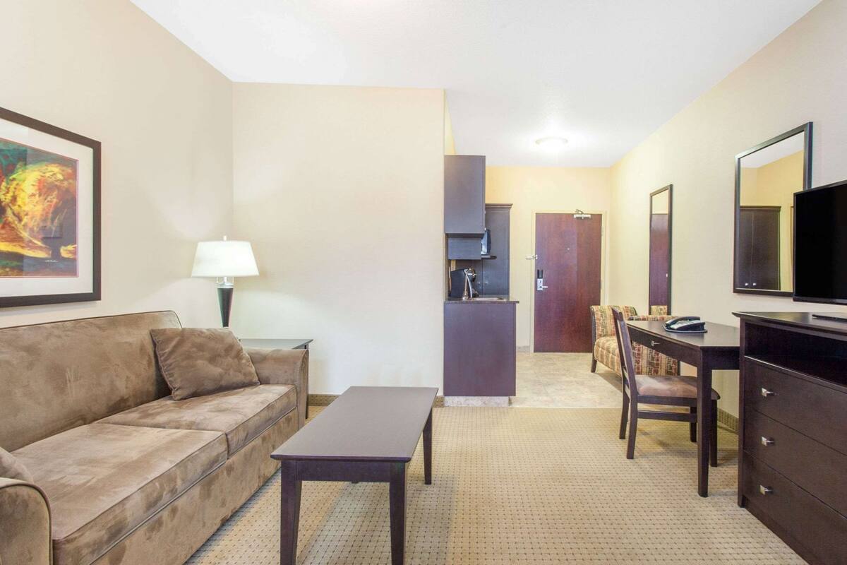 Habitación de hotel con sofá, una mesita, escritorio con silla, televisor, espejo y puerta que da a un baño.