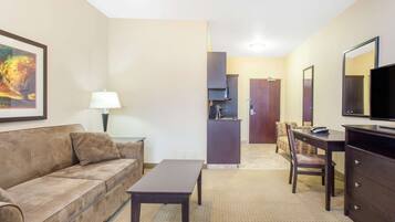 Une chambre d’hôtel équipée d’un canapé, d’une table, d’un téléviseur, d’un bureau, d’une chaise, d’une lampe, d’un coin cuisine et d’un tableau encadré au mur.