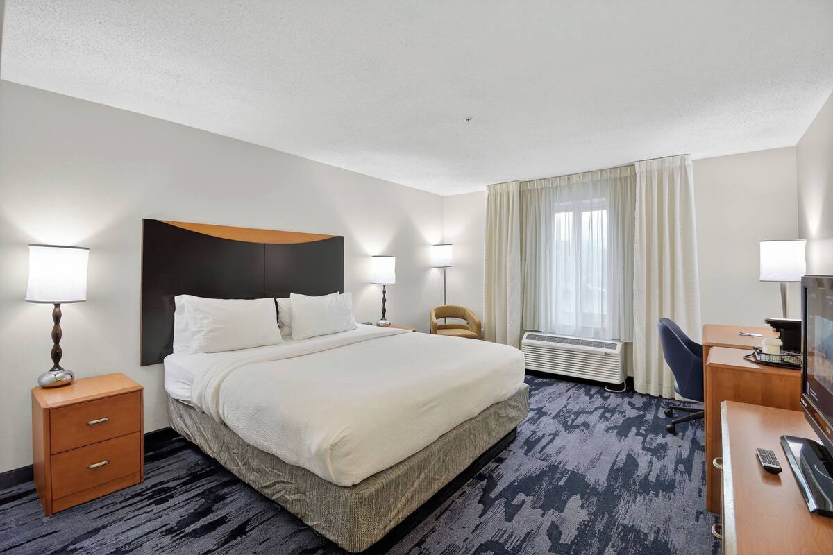 Quarto de hotel com cama, abajures, escrivaninha com televisão e uma cadeira.