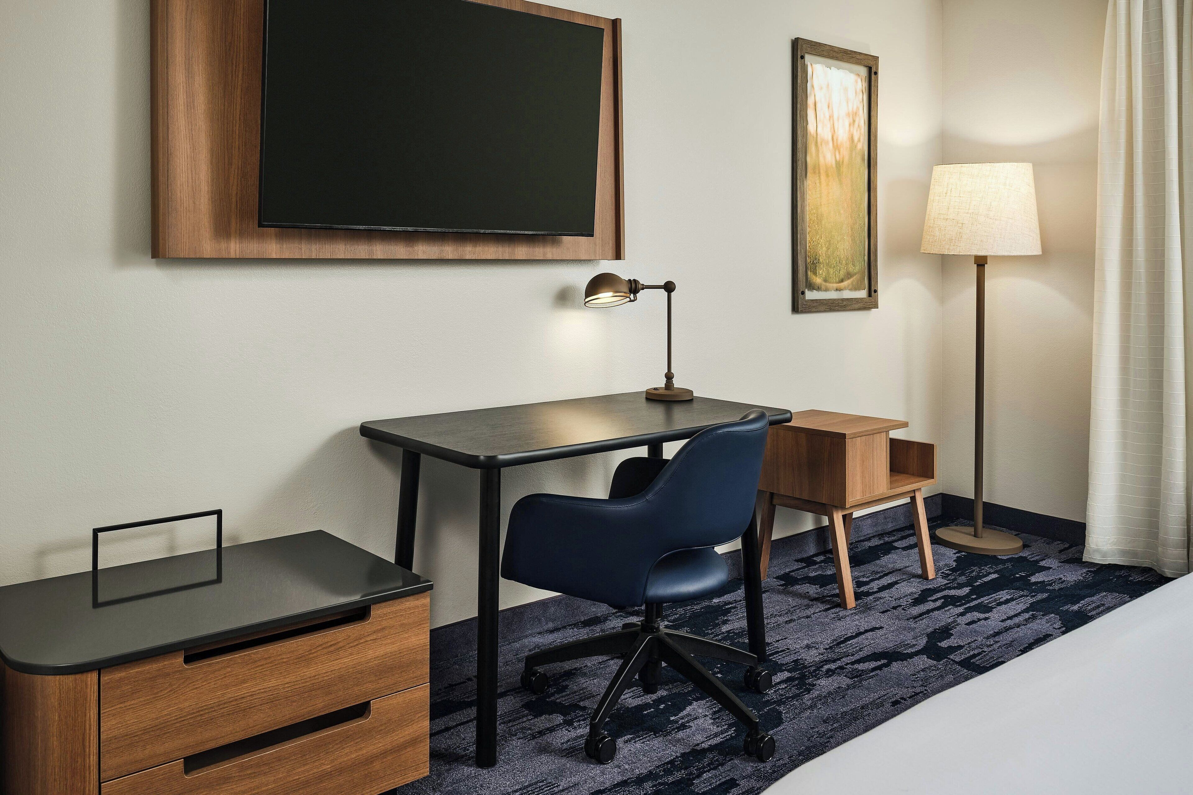 A hotel room with a desk, chair, bedside table, lamp, and artwork on the wall.