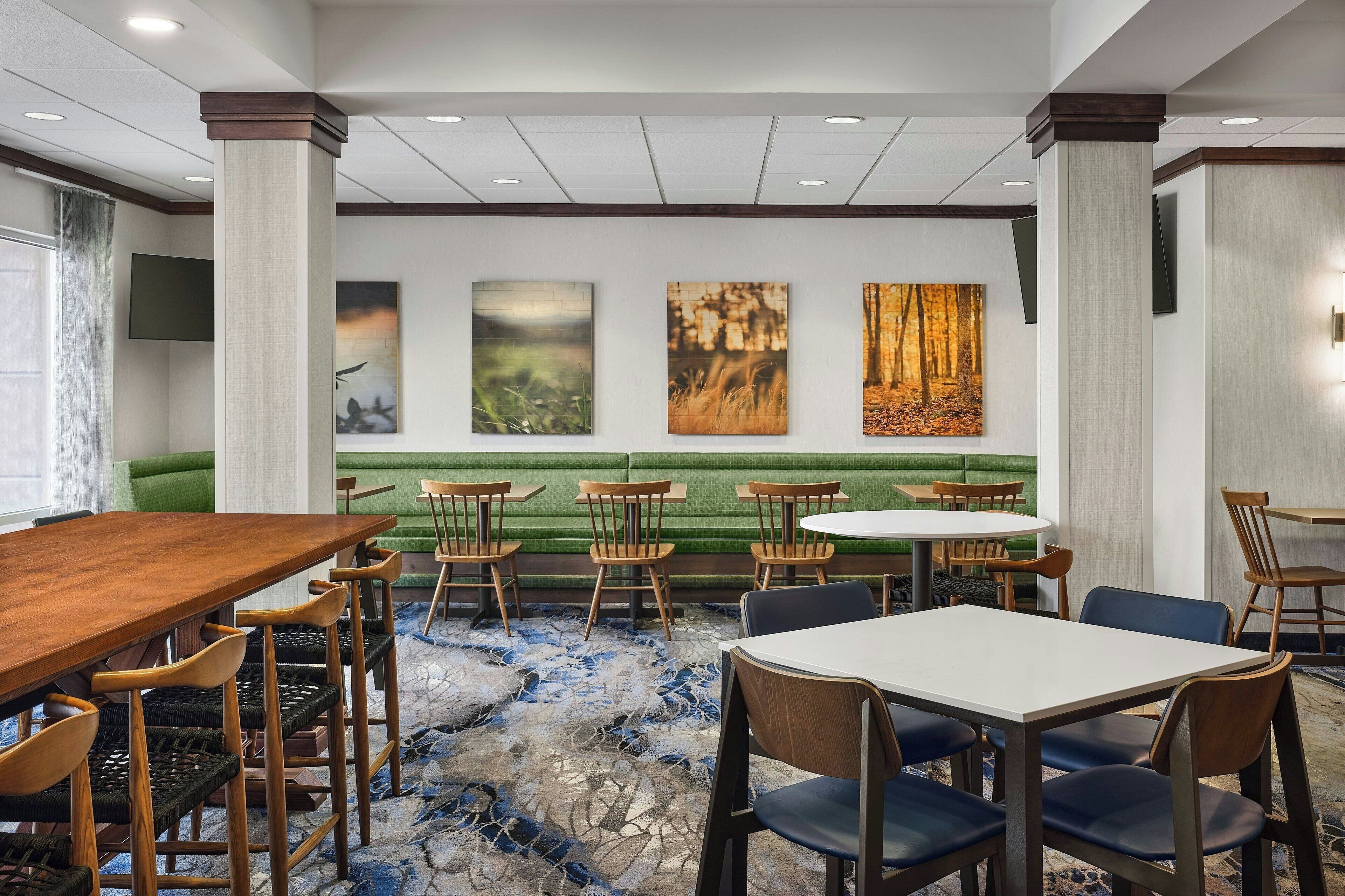 A hotel dining area with a long wooden table, several round tables, and a green sofa.