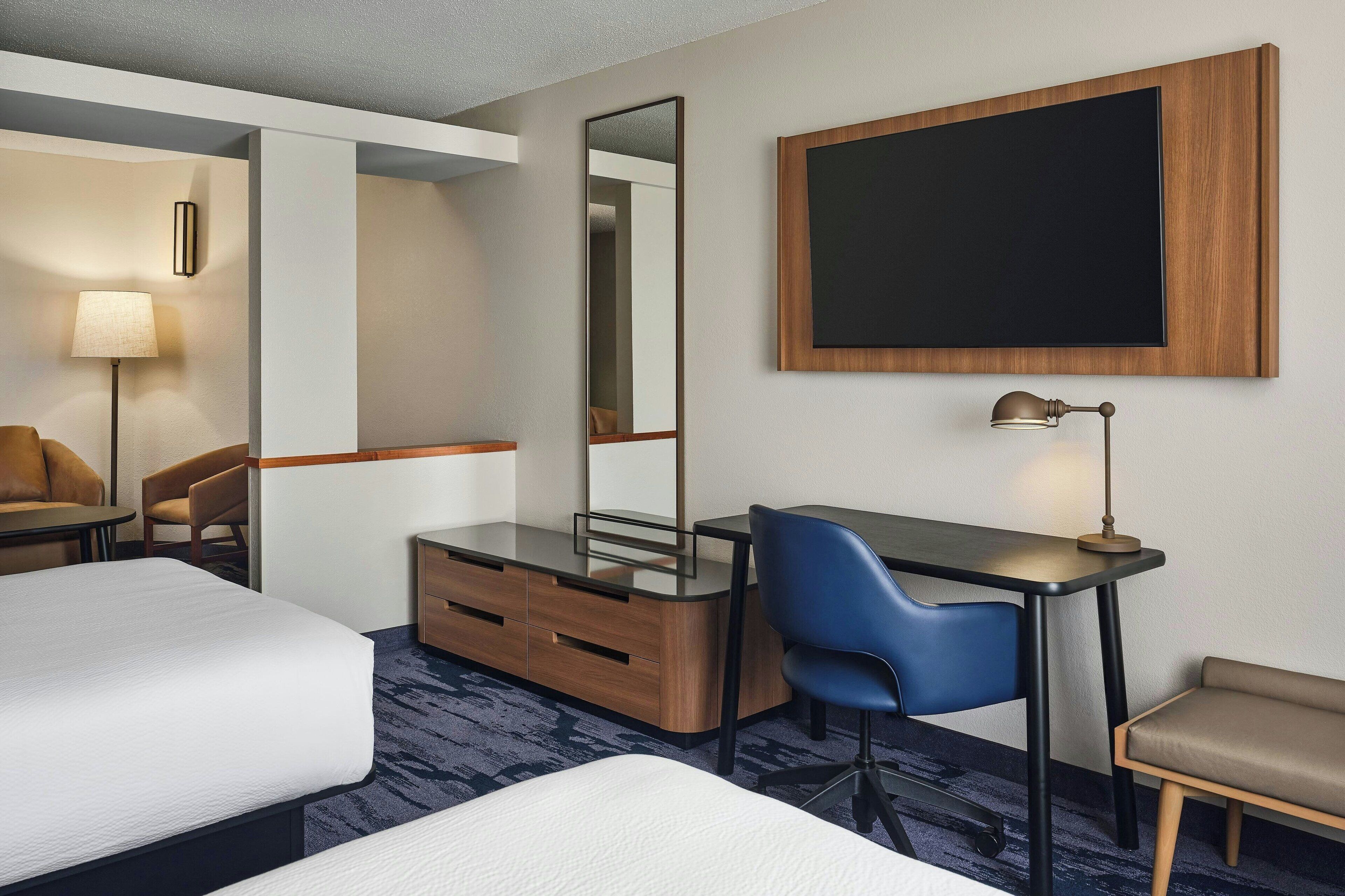 Habitación de hotel con cama, escritorio con silla, cómoda, espejo, lámpara y un televisor grande montado en la pared.