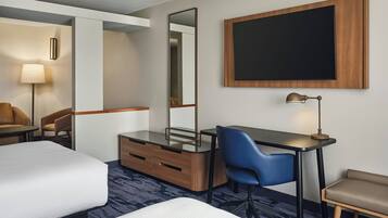 Habitación de hotel con cama, escritorio con silla, cómoda, espejo, lámpara y un televisor grande montado en la pared.