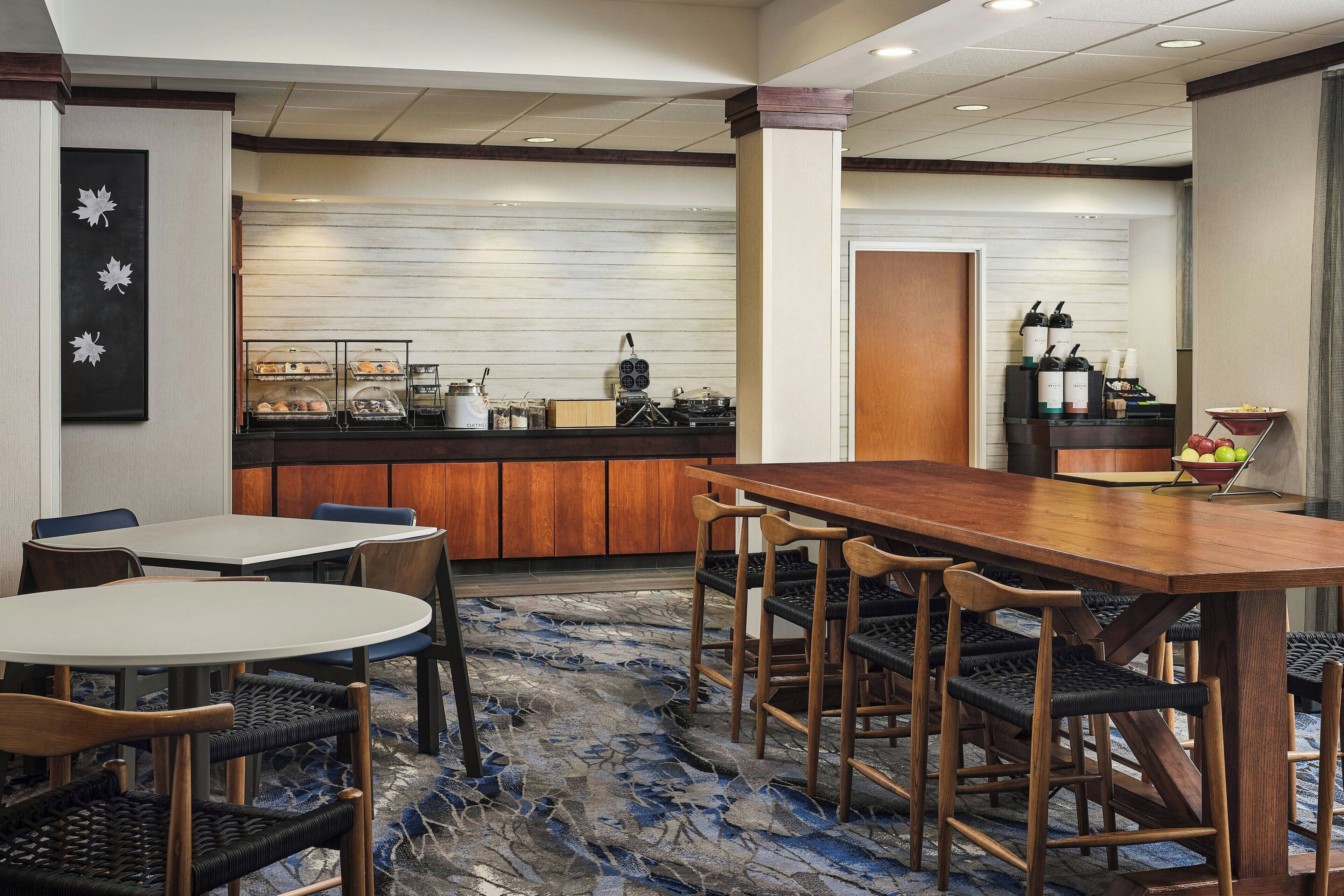 A hotel dining area with a long wooden table and chairs, a buffet counter, and a wall-mounted wine cooler.