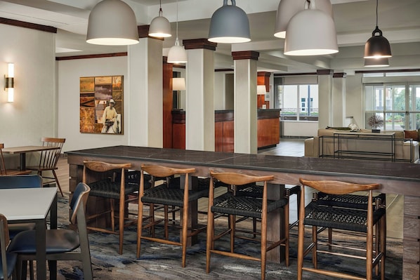 A modern hotel lobby with a long bar area, wooden chairs, and pendant lighting.