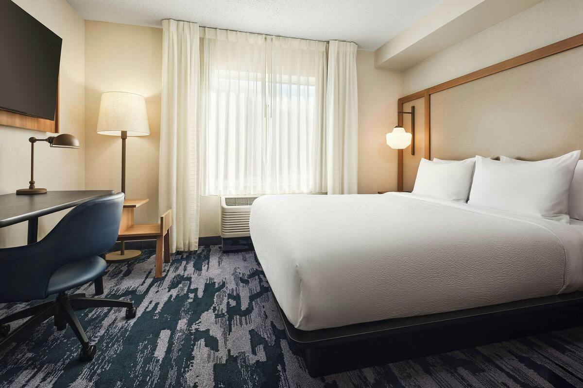 A hotel room with a bed, desk, chair, and a large window with curtains.