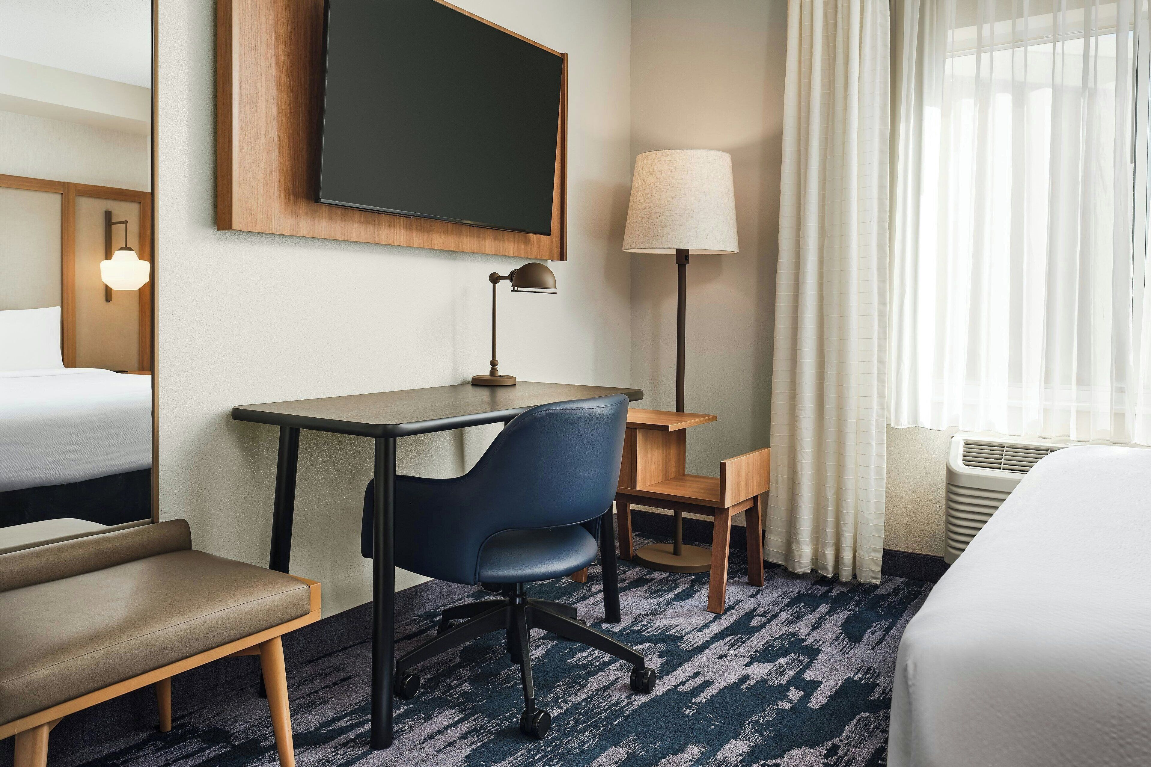 A hotel room with a bed, a desk with a chair, a television, a lamp, and a window with curtains.