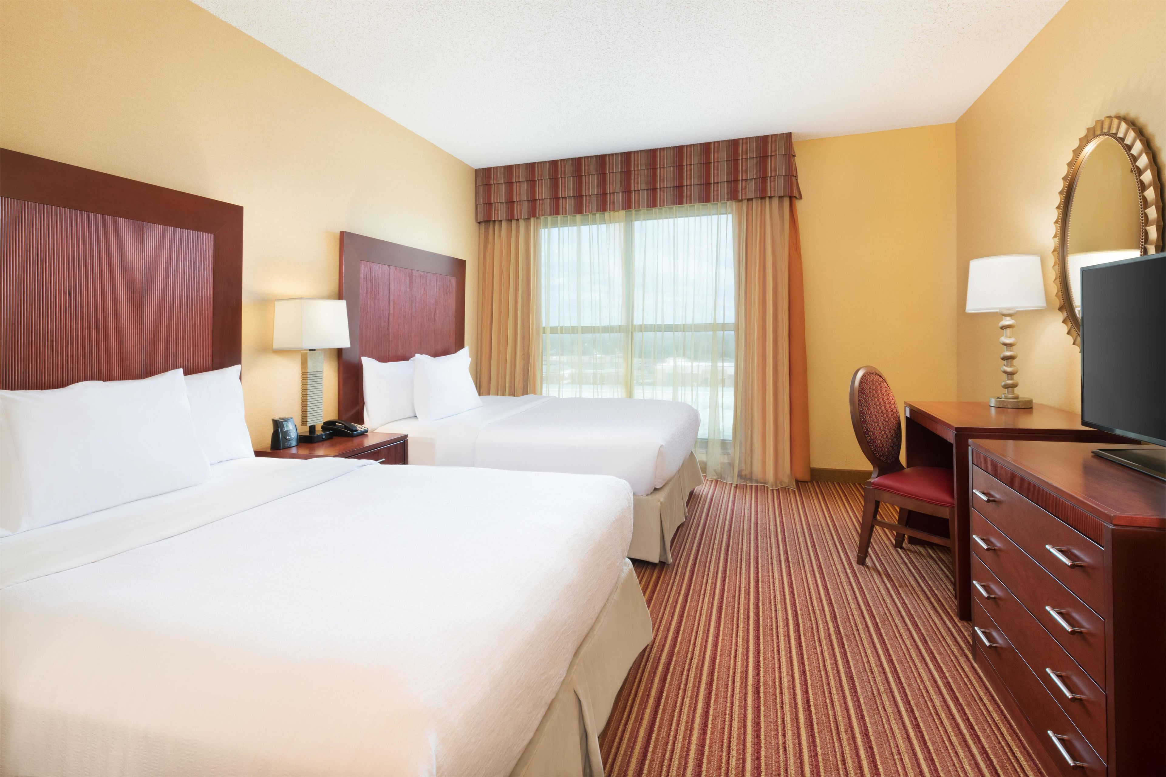A hotel room with two beds, a desk with a chair, a television, and a large window with curtains.