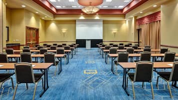 Un salón de conferencias con filas de mesas y sillas, una pantalla de proyector y suelo alfombrado.
