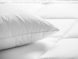 A close-up of a white bed with a neatly arranged pillow and a smooth, unadorned headboard.