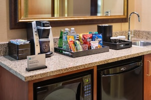 Cocina de habitación de hotel con cafetera, botellas de agua, aperitivos y una taza de café.