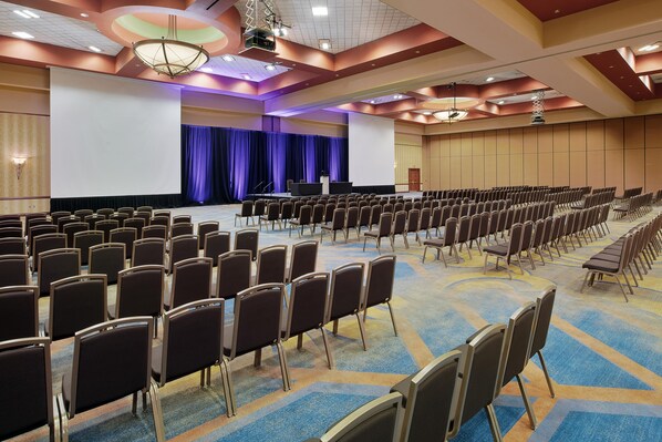 Un salón de conferencias con filas de sillas, un escenario con un atril y una pantalla grande.