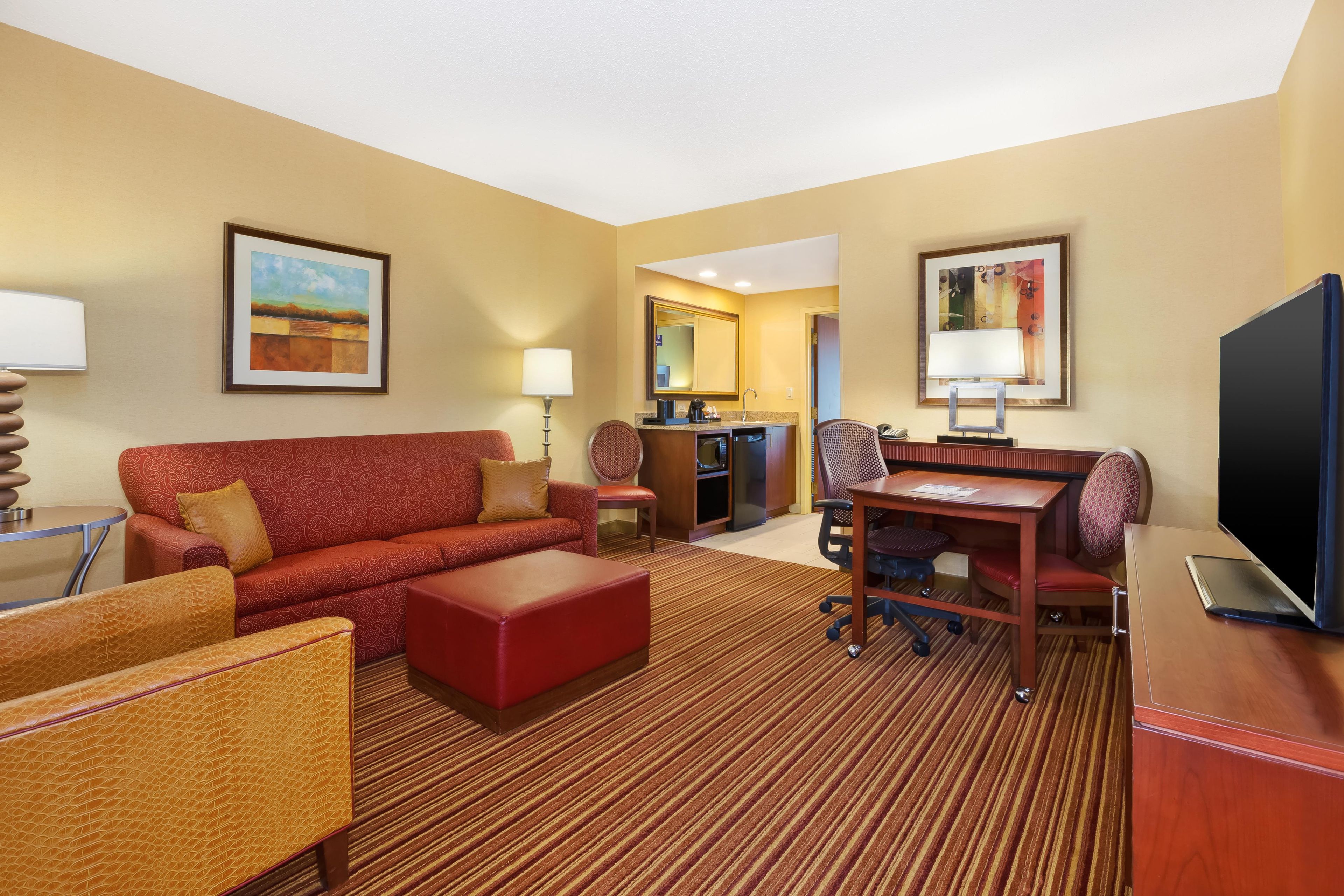 Habitación de hotel con sofá, sillón, reposapiés, mesa de comedor, sillas, televisor, escritorio con lámpara y obras de arte en las paredes.