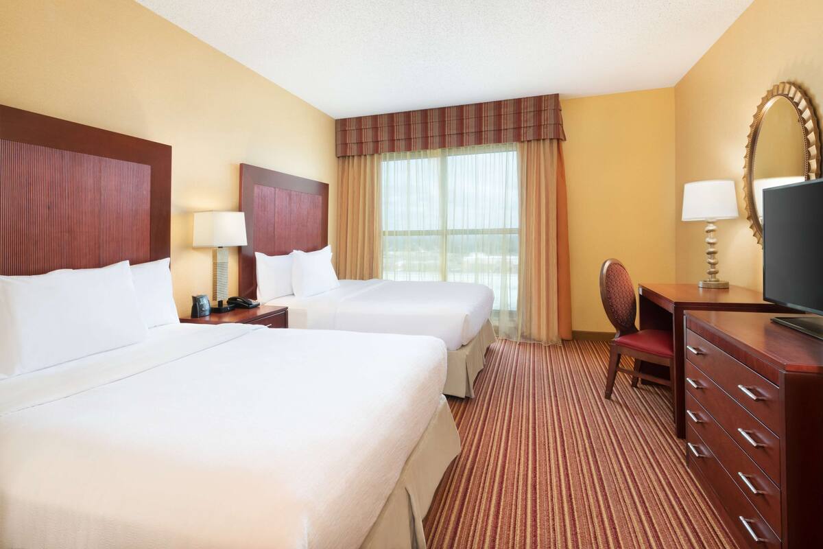 A hotel room with two beds, a desk, a TV, and a large window with curtains.