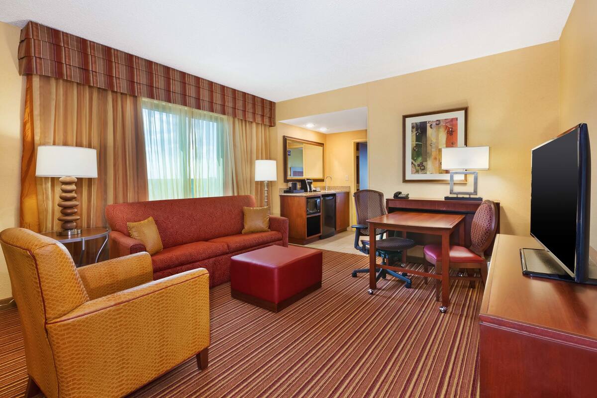 A hotel room with a red sofa, a red ottoman, a wooden desk with a chair, a television, and a large window with curtains.