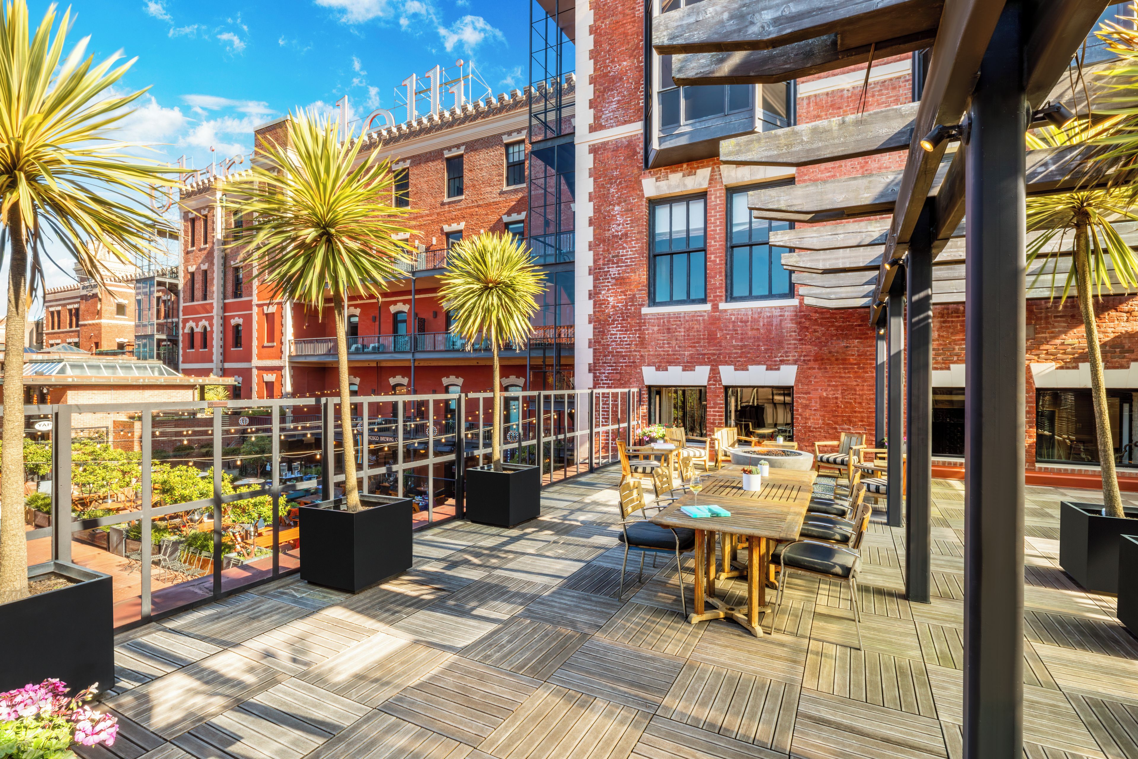 A rooftop terrace with outdoor seating, a brick building in the background, and palm trees.