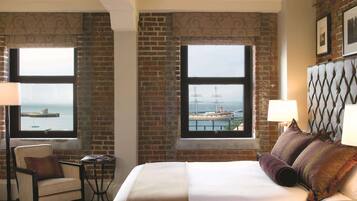 A hotel room with a bed, a chair, a table, and two windows with views of a dock and a ship.