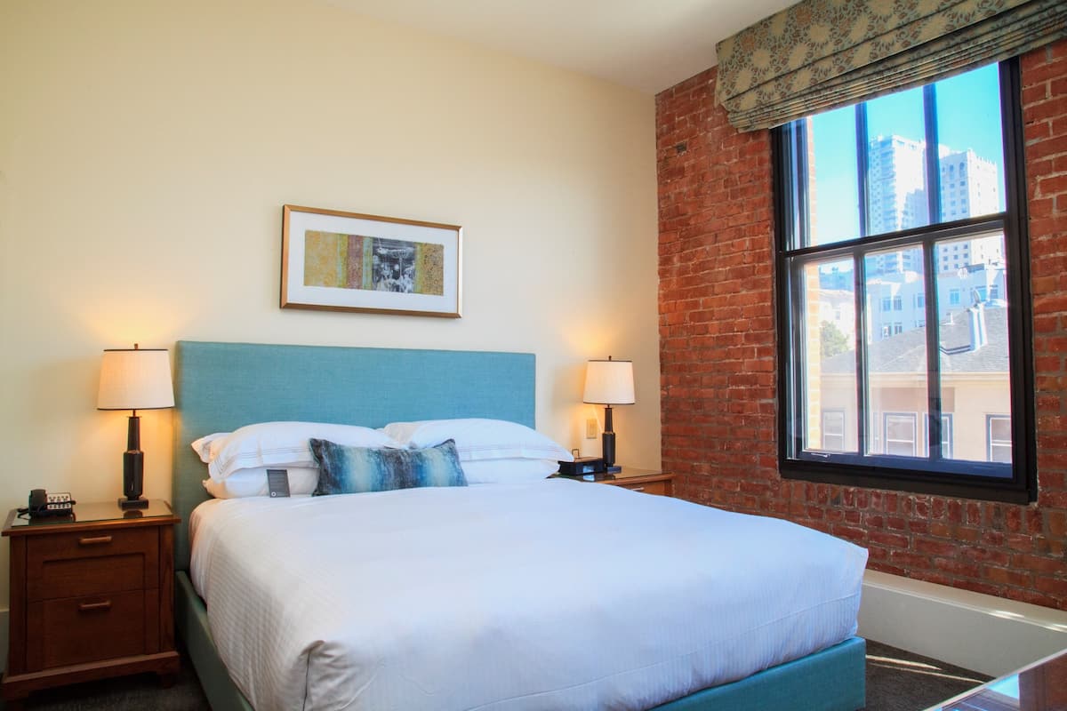 A hotel room with a large bed, two bedside tables with lamps, a framed picture on the wall, and a large window with a view of buildings.