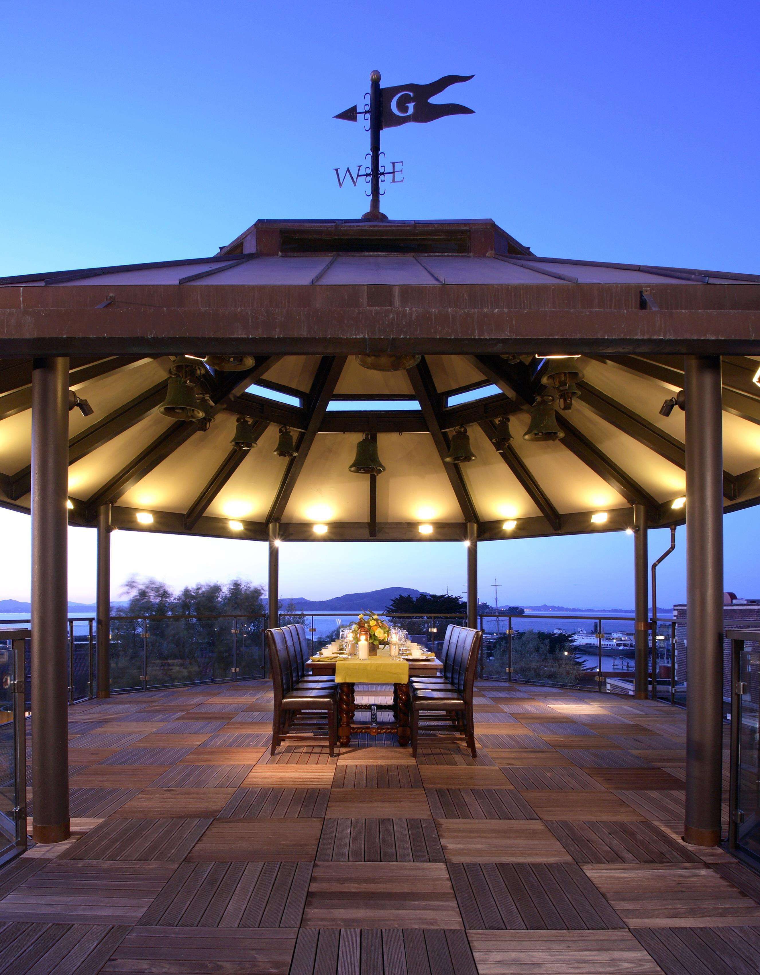 A covered outdoor dining area with a table set for two, surrounded by chairs, under a metal roof with a weather vane.