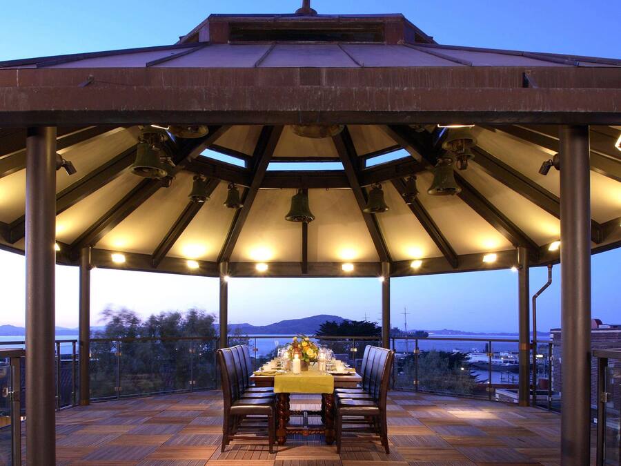 Un área de comedor al aire libre techada con una mesa lista para dos personas, rodeada de sillas, bajo un techo de metal con veleta.