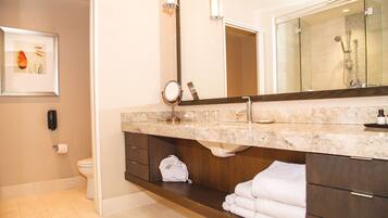 A modern bathroom with a marble countertop, a large mirror, a glass shower enclosure, and a neatly folded towel on a shelf.
