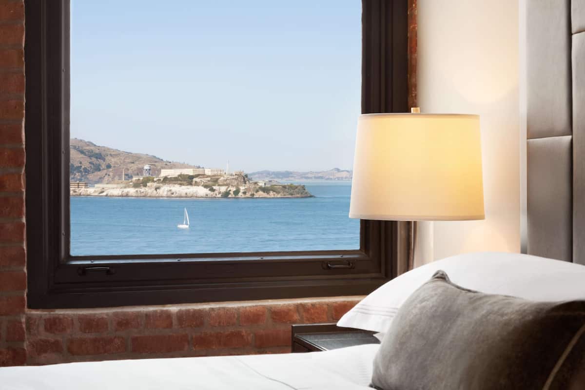 A hotel room with a large window offering a view of a coastal area, a bed, and a bedside lamp.