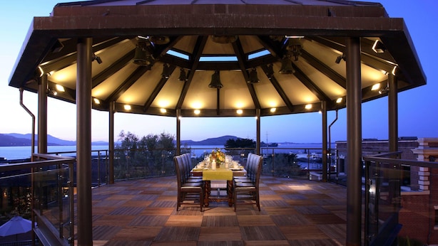 A covered outdoor dining area with a wooden deck, a table set for two, and a view of the sea and mountains in the background. - Fairmont Heritage Place, Ghirardelli Square (San Francisco)