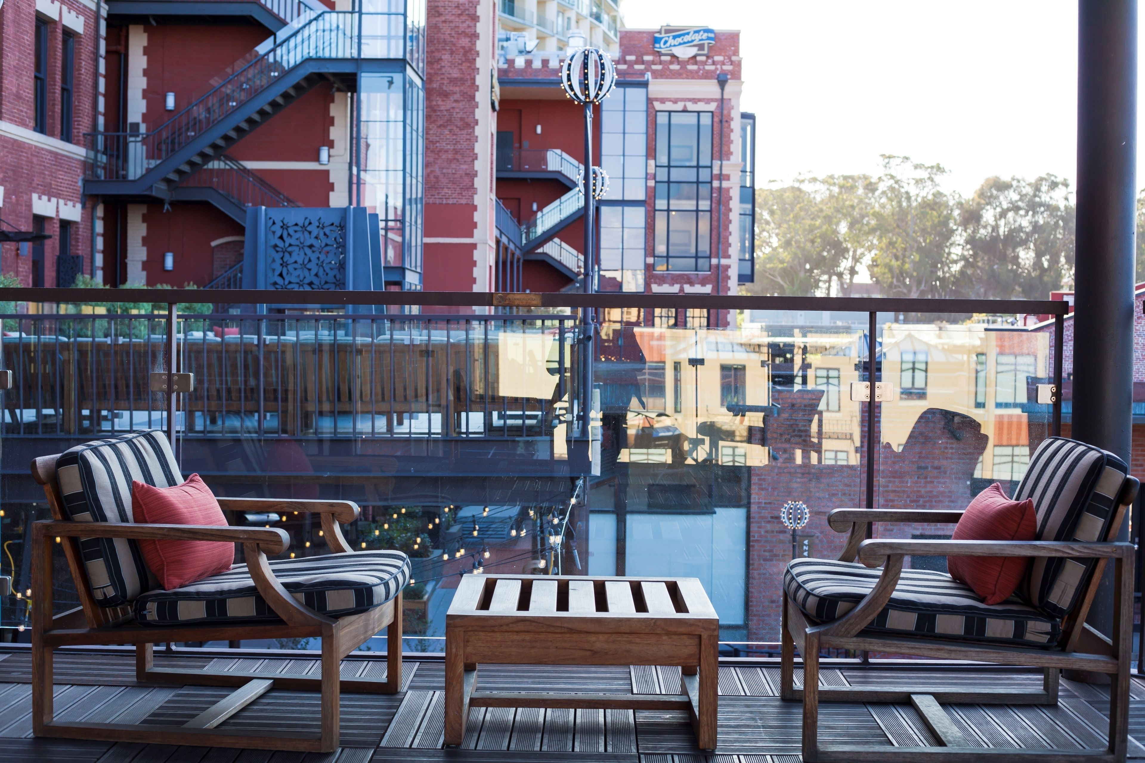 A balcony with two striped chairs and a wooden table, overlooking a cityscape with brick buildings and a staircase.