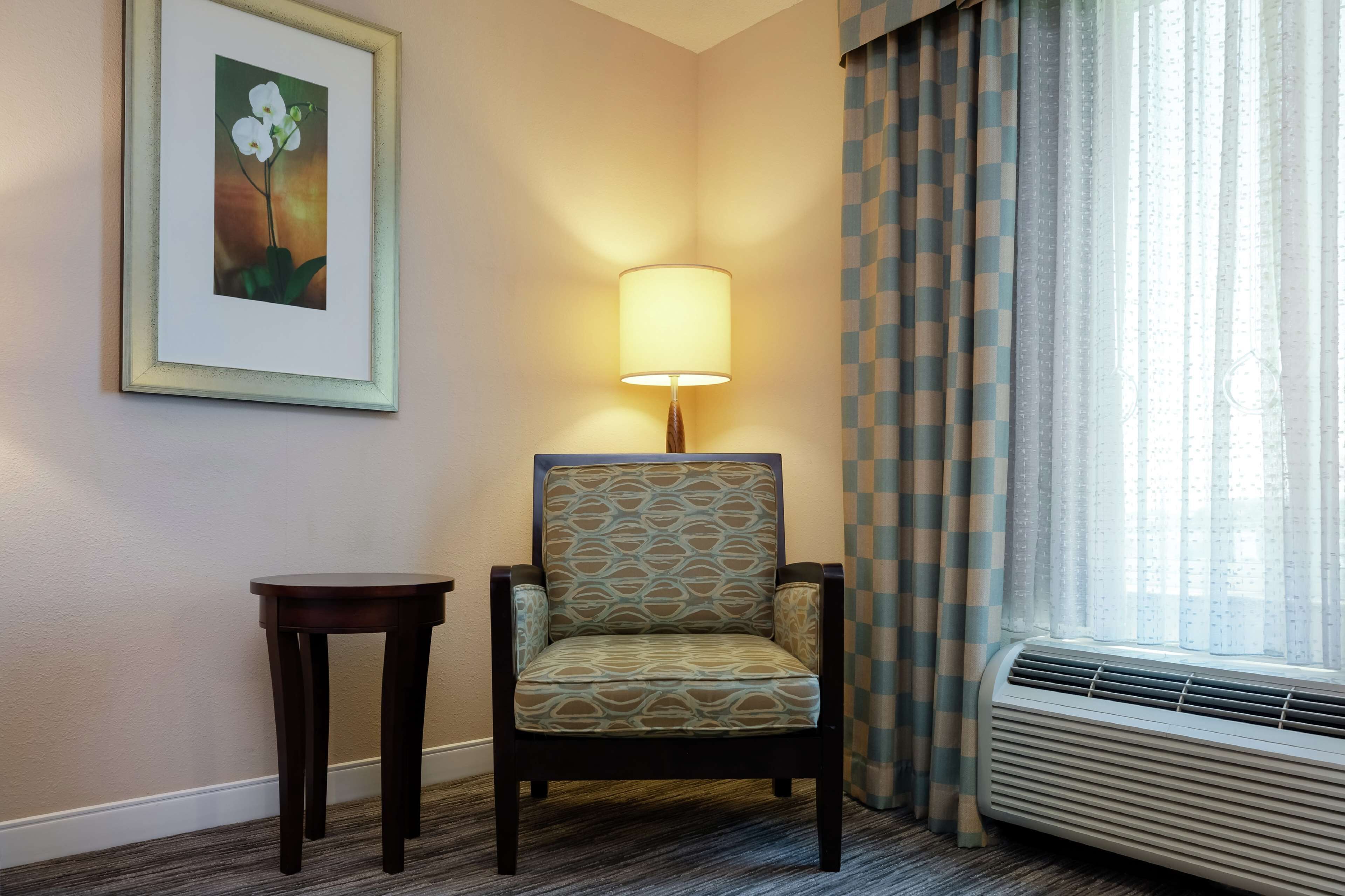 Une chambre d’hôtel avec un fauteuil à motifs, une petite table, un tableau encadré au mur et une fenêtre avec des rideaux.