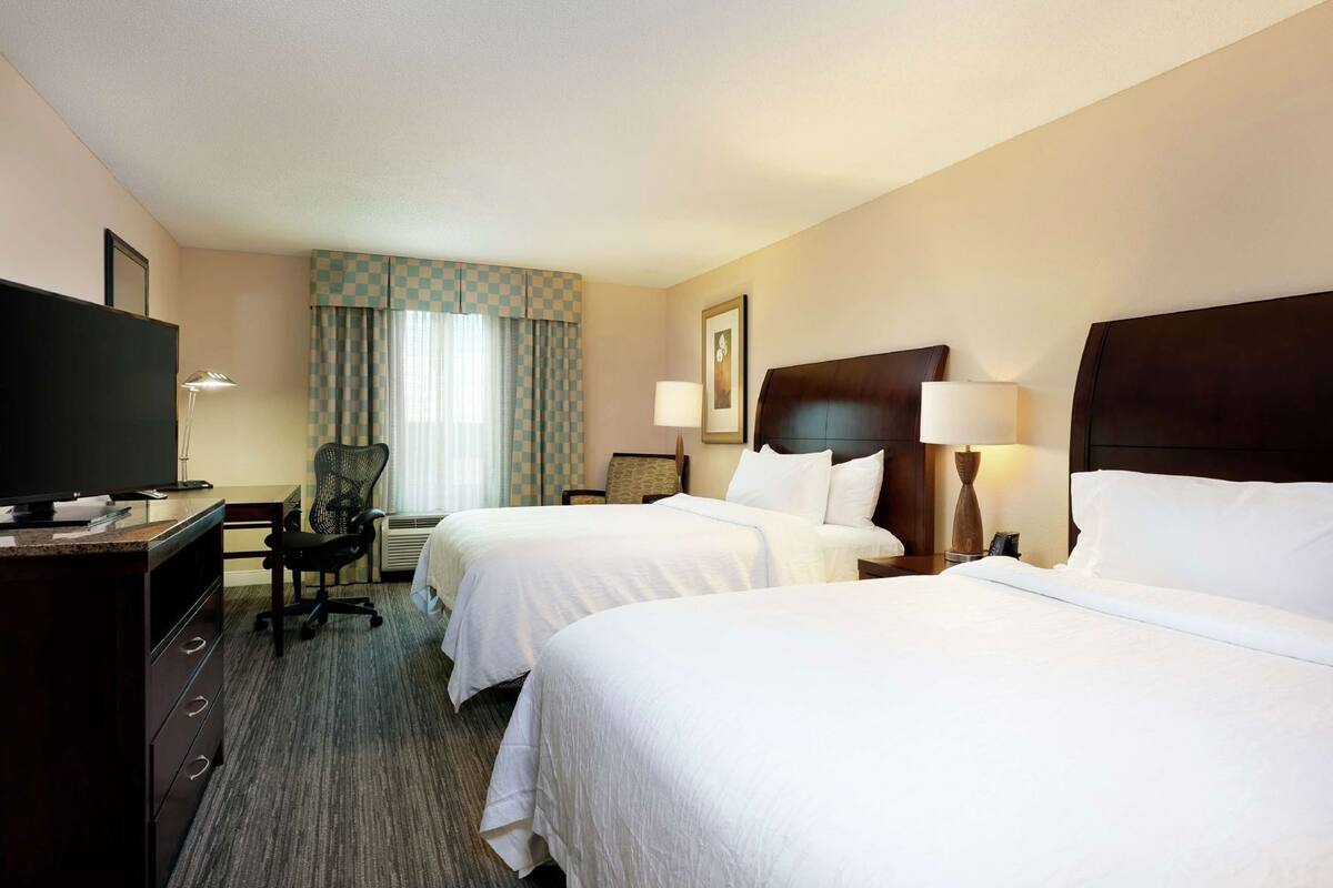 Een hotelkamer met twee bedden, een bureau met een televisie, een stoel en een raam met gordijnen.