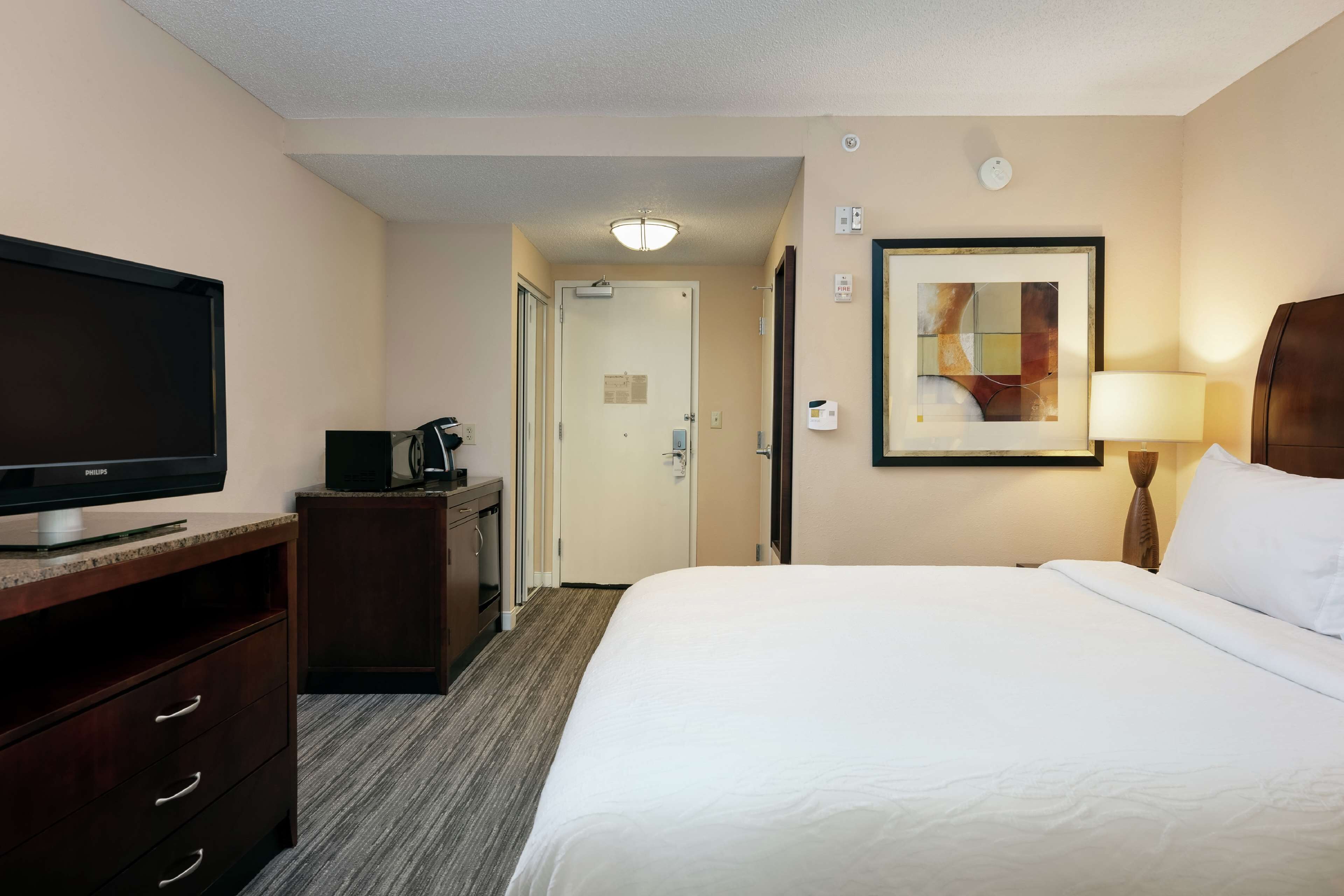A hotel room with a bed, a television, a desk with a coffee maker, a framed picture on the wall, and a lamp.