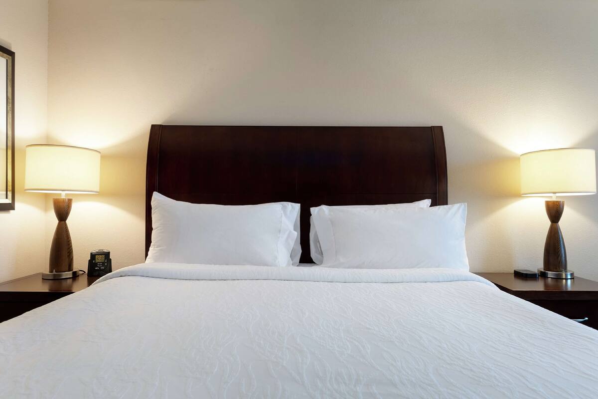 Een netjes opgemaakt bed met witte lakens, twee nachtkastjes met lampen en een donker hoofdbord.