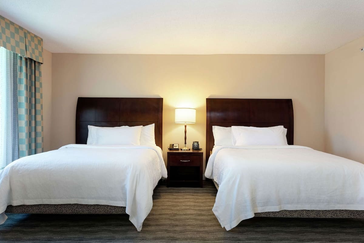 A hotel room with two beds, a nightstand with a lamp, and a window with curtains.