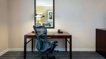 Quarto de hotel com uma secretária de madeira, uma cadeira de escritório preta, uma luminária e um quadro na parede.