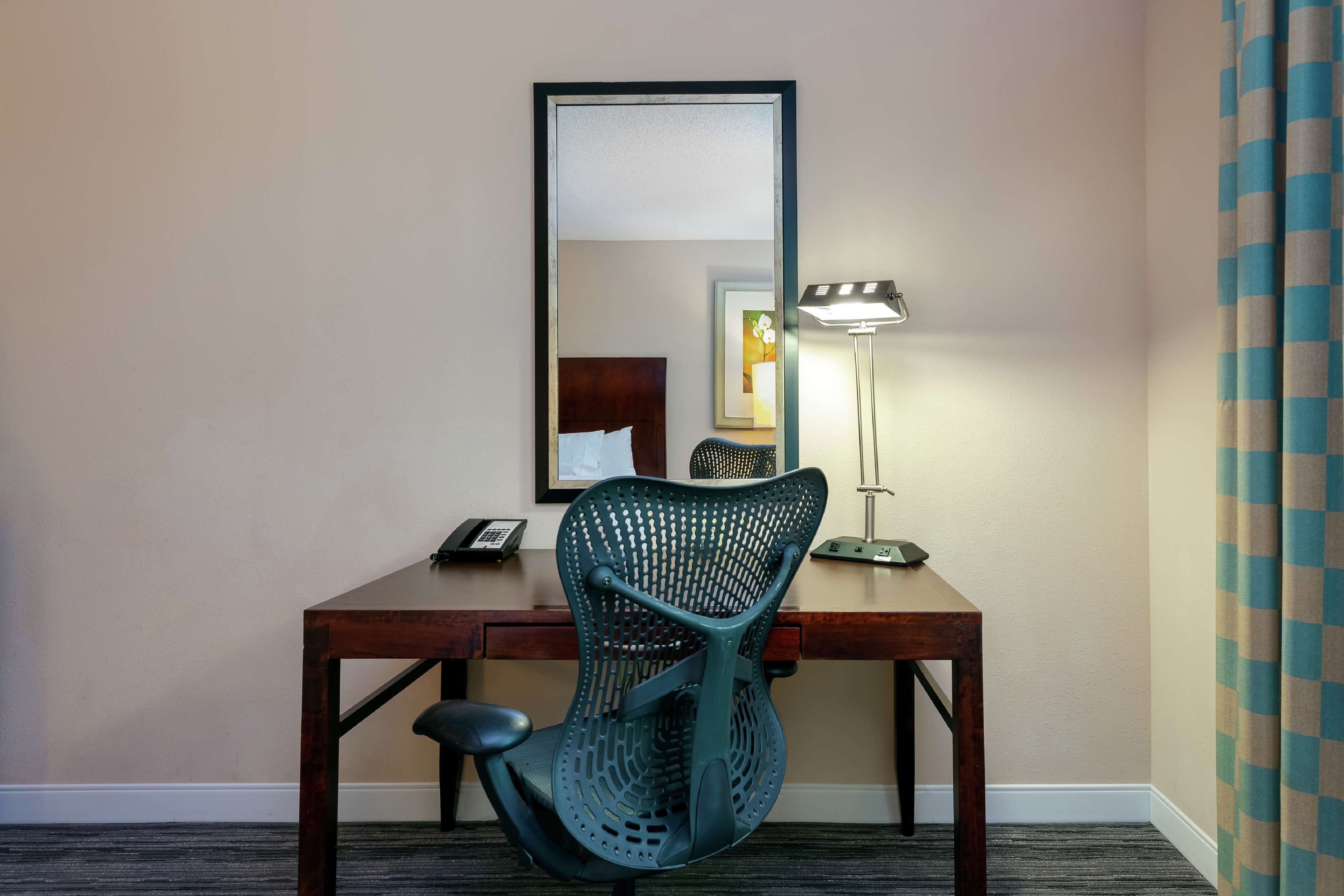 A hotel room with a wooden desk, a green mesh chair, a desk lamp, and a mirror.