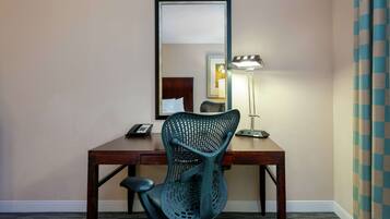 A hotel room with a wooden desk, a green mesh chair, a desk lamp, and a mirror.