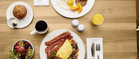 Uma mesa de café da manhã com pratos de ovos, bacon, torradas e frutas. Há também xícaras de café e suco, e um muffin.