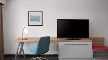Quarto de hotel moderno com escrivaninha de madeira, cadeira de escritório azul, luminária de chão, TV de tela plana e um pequeno armário com assento vermelho.