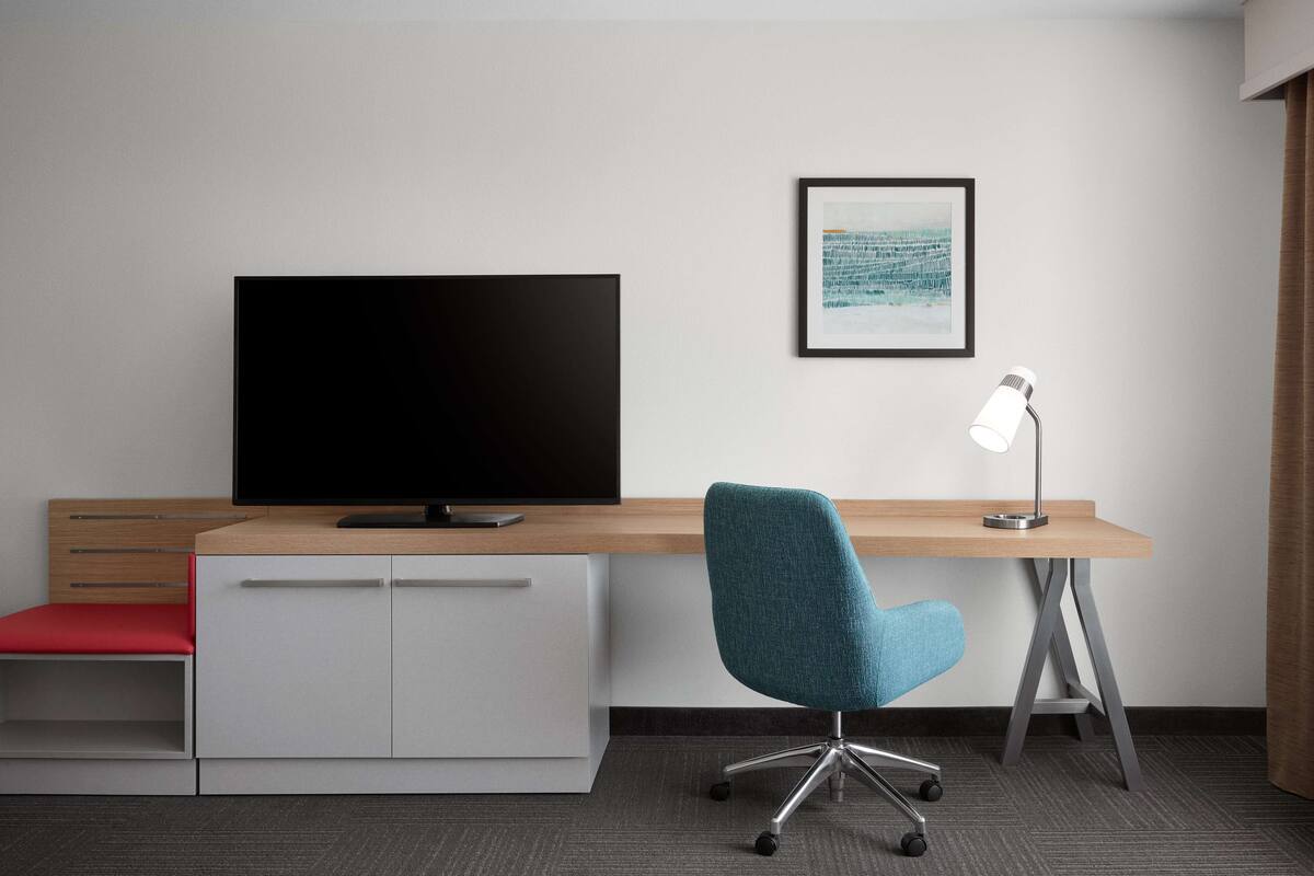 A modern hotel room with a flat-screen TV, a desk with a chair, a framed picture on the wall, and a desk lamp.
