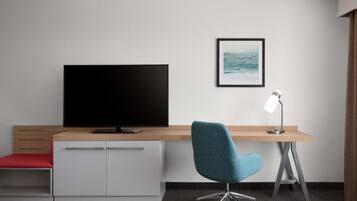 Et moderne hotelværelse med et fladskærms-TV, et skrivebord med stol, et indrammet billede på væggen og en skrivebordslampe.