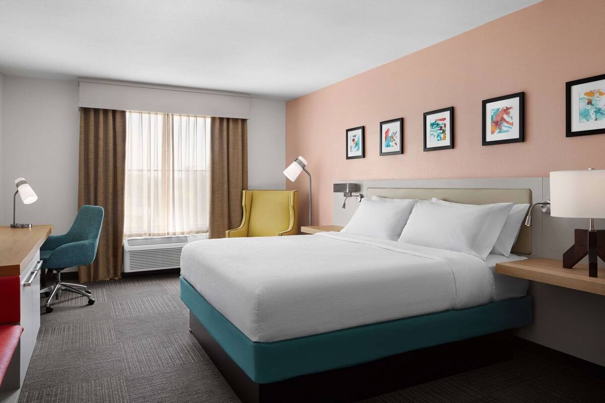 A hotel room with a bed, desk, chair, and artwork on the wall.