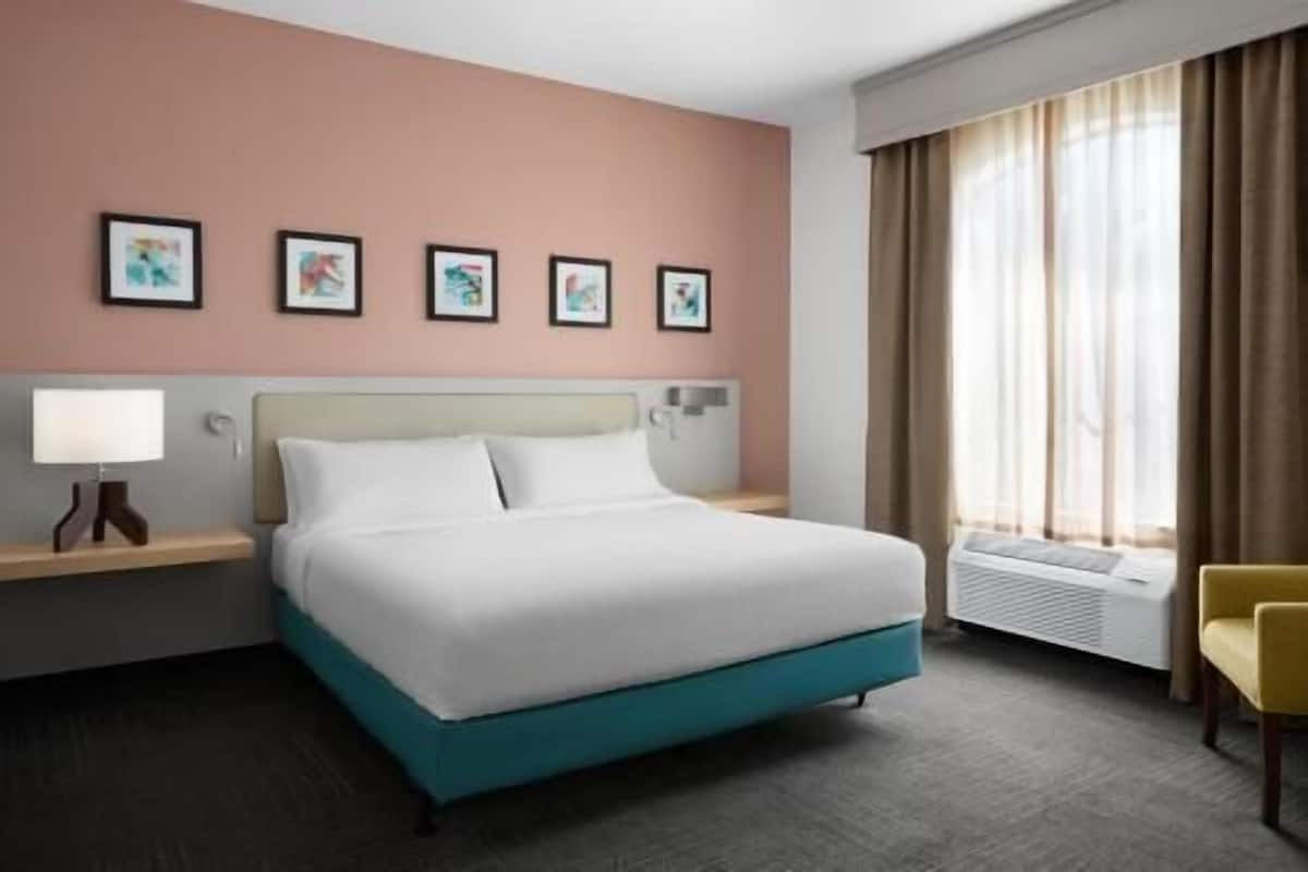 A hotel room with a bed, a nightstand, a lamp, a chair, and a window with curtains.