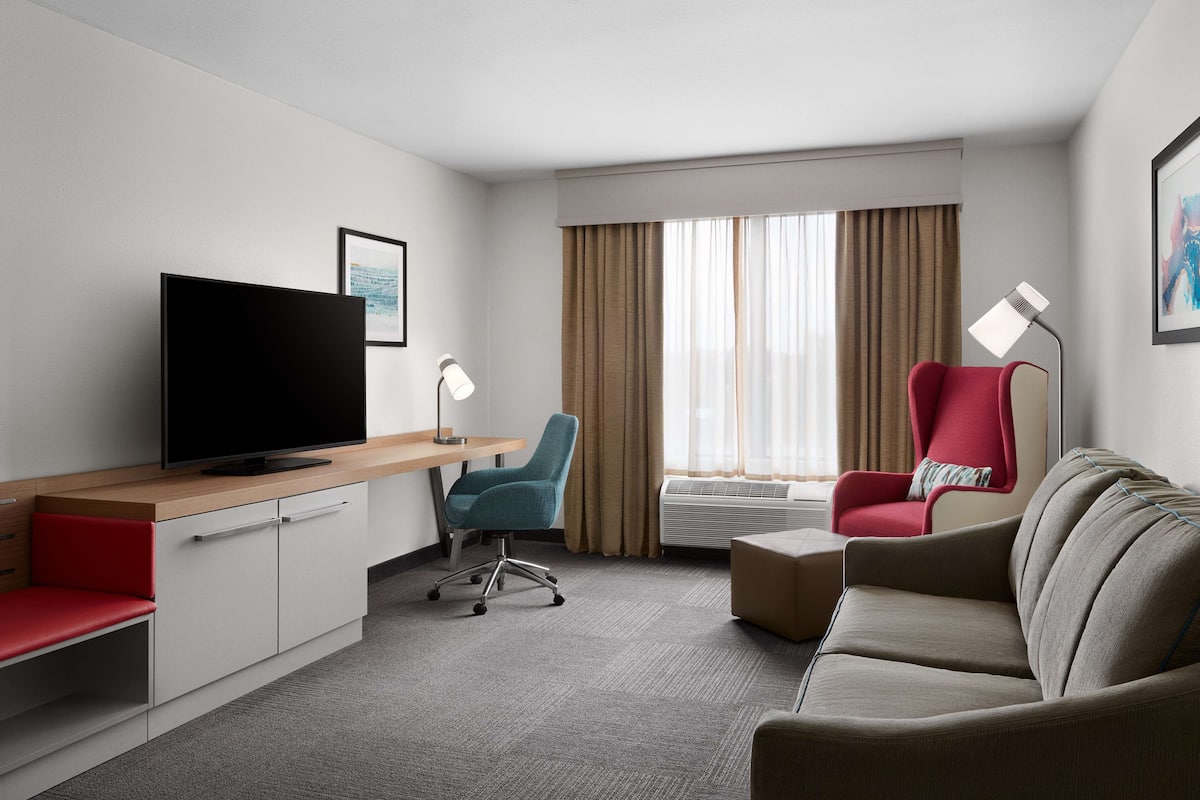 A hotel room with a flat-screen TV, a desk, a red armchair, a beige sofa, and a large window with curtains.